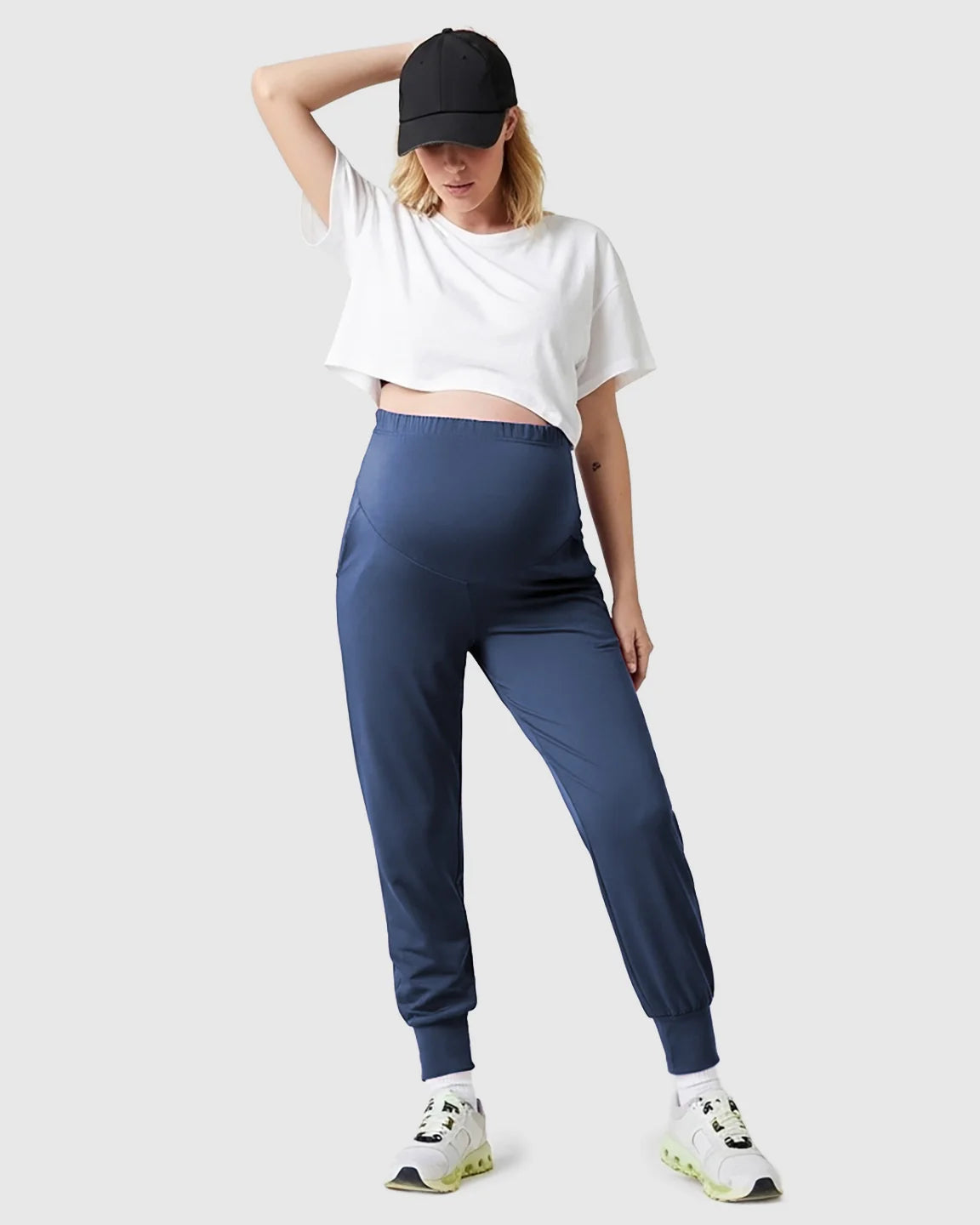 Woman in a black cap, white crop top, and supportive navy blue maternity joggers with a high-waist design.