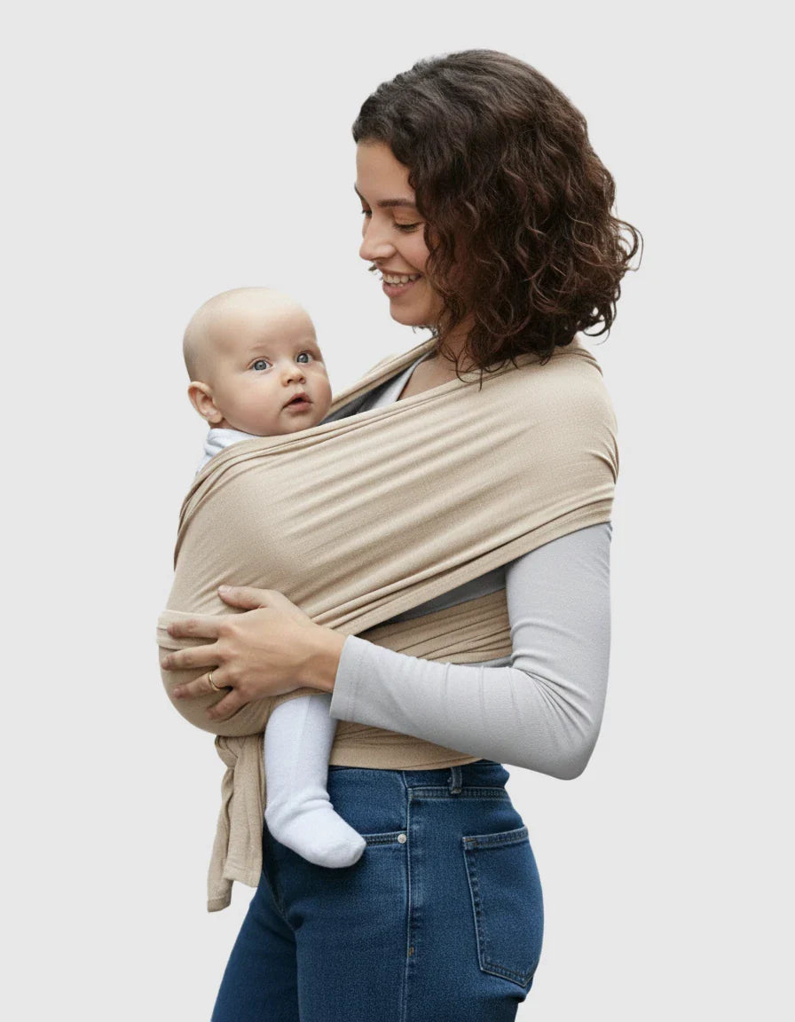 Smiling woman wearing a beige adjustable wrap-around baby carrier with a baby nestled inside.