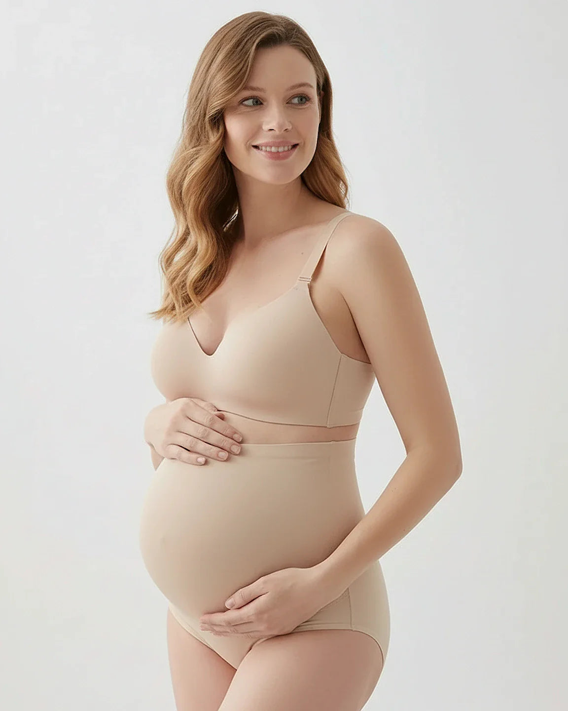 Smiling pregnant woman in a beige seamless maternity bra and high-waist briefs, gently holding her belly.