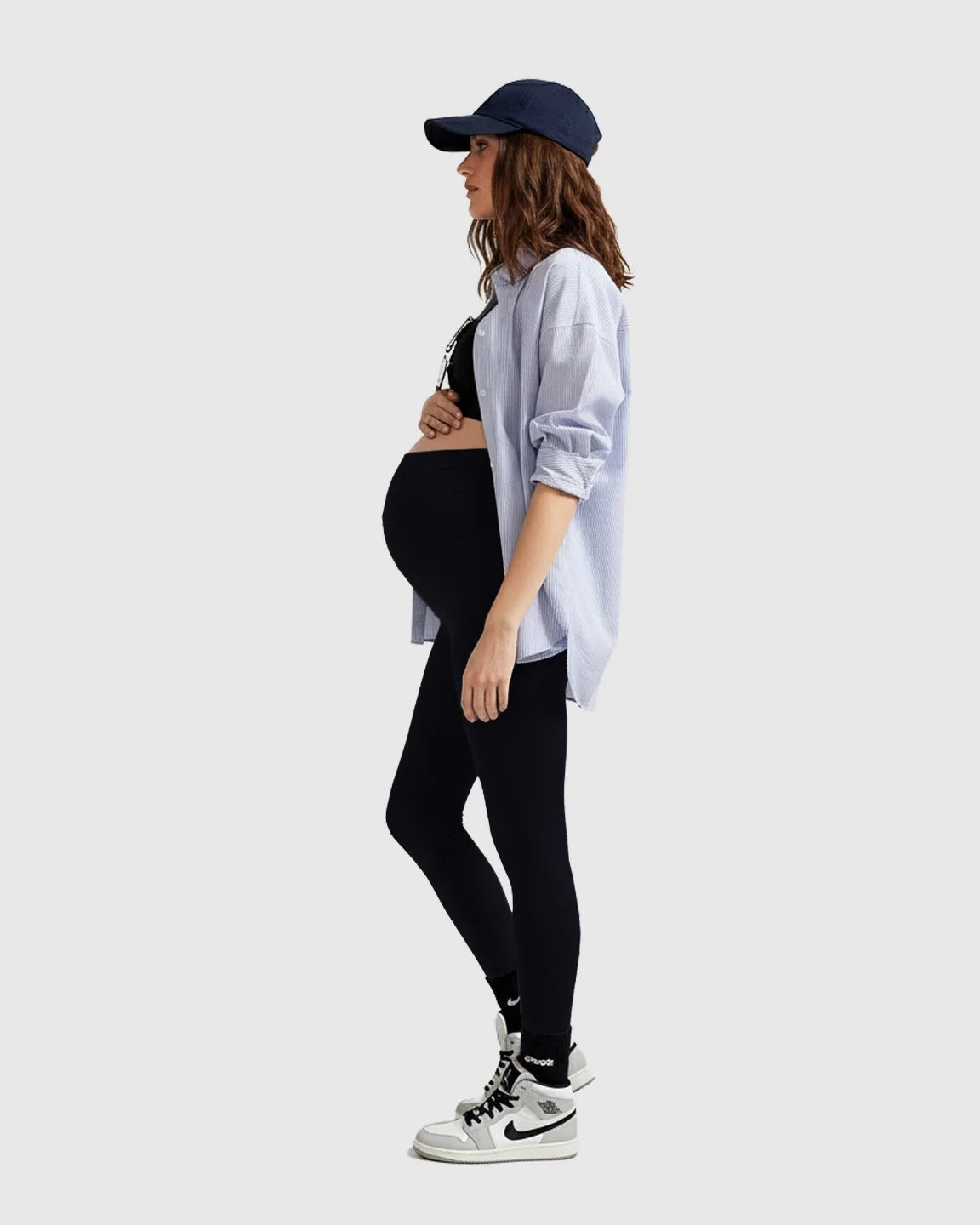 Pregnant woman in black midi maternity leggings, a striped shirt, and a navy baseball cap.