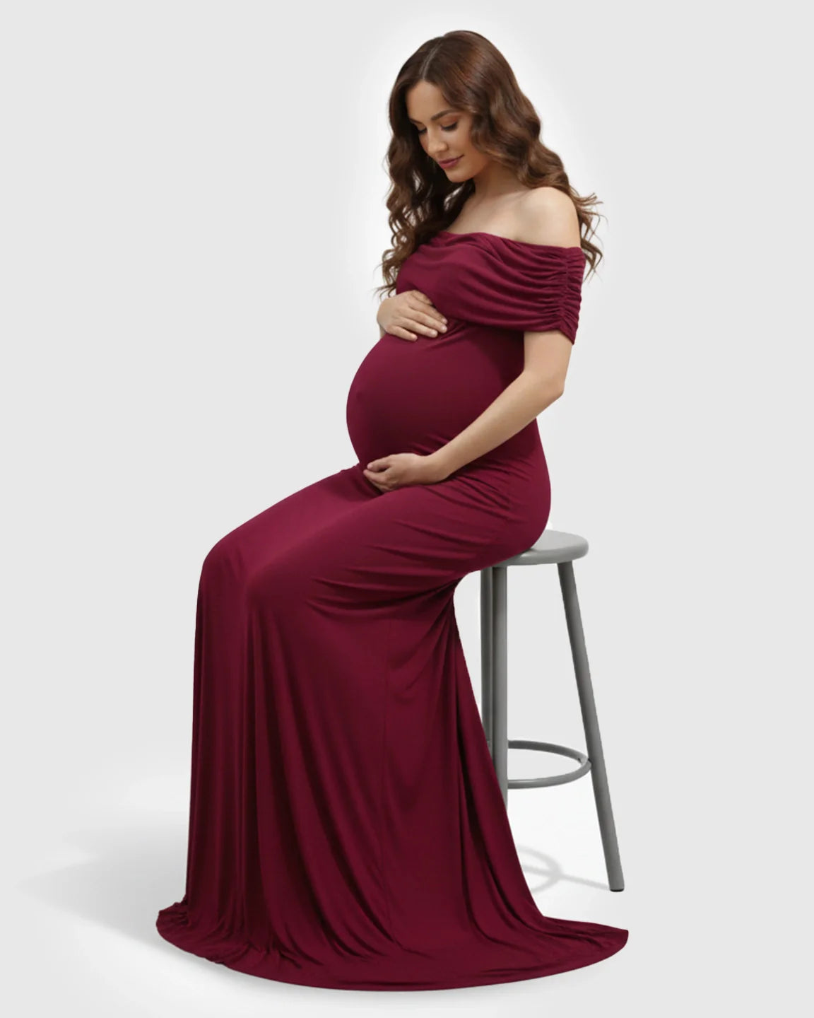 Maternity model in a deep red off-shoulder bodycon mermaid gown, cradling her bump while seated.
