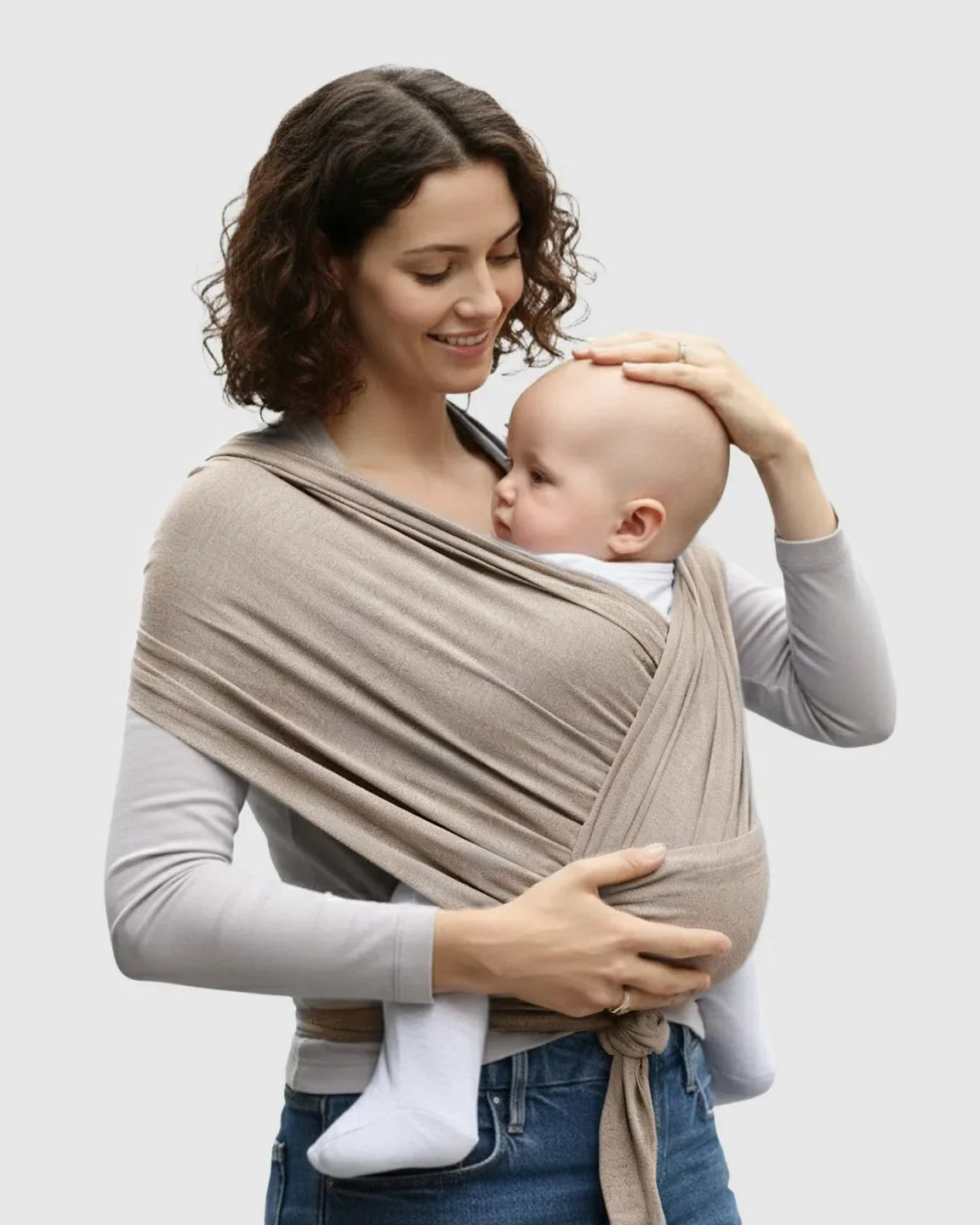 Woman smiling, wearing a soft, beige adjustable wrap-around baby carrier with a baby snuggled close.