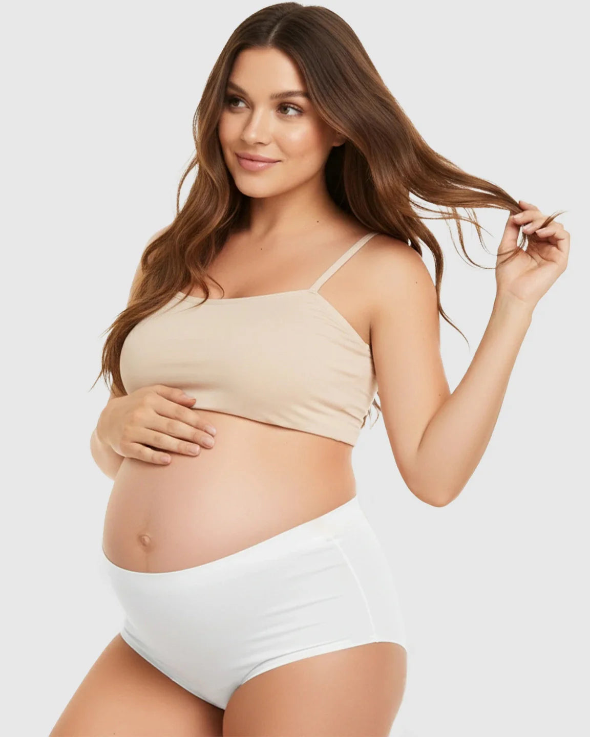 Pregnant woman in a beige seamless maternity bra and white high-waist support underwear, gently holding her belly.
