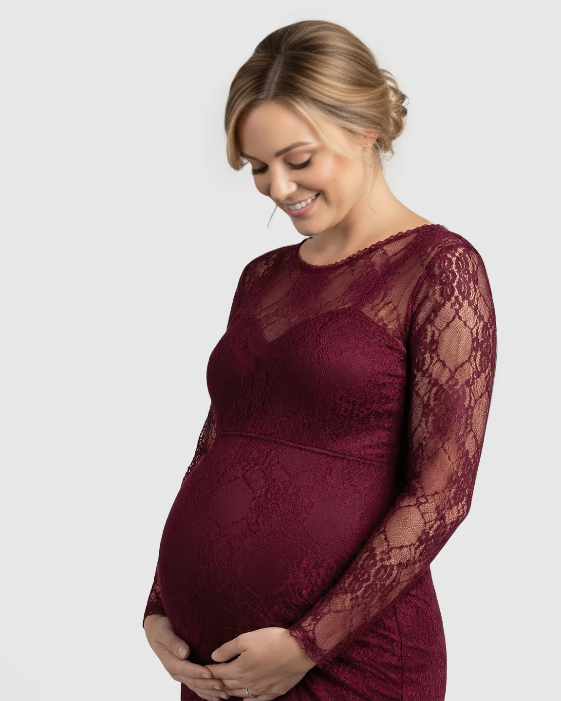 Smiling pregnant woman in a long-sleeved sheer lace trumpet maternity gown in a deep burgundy color.