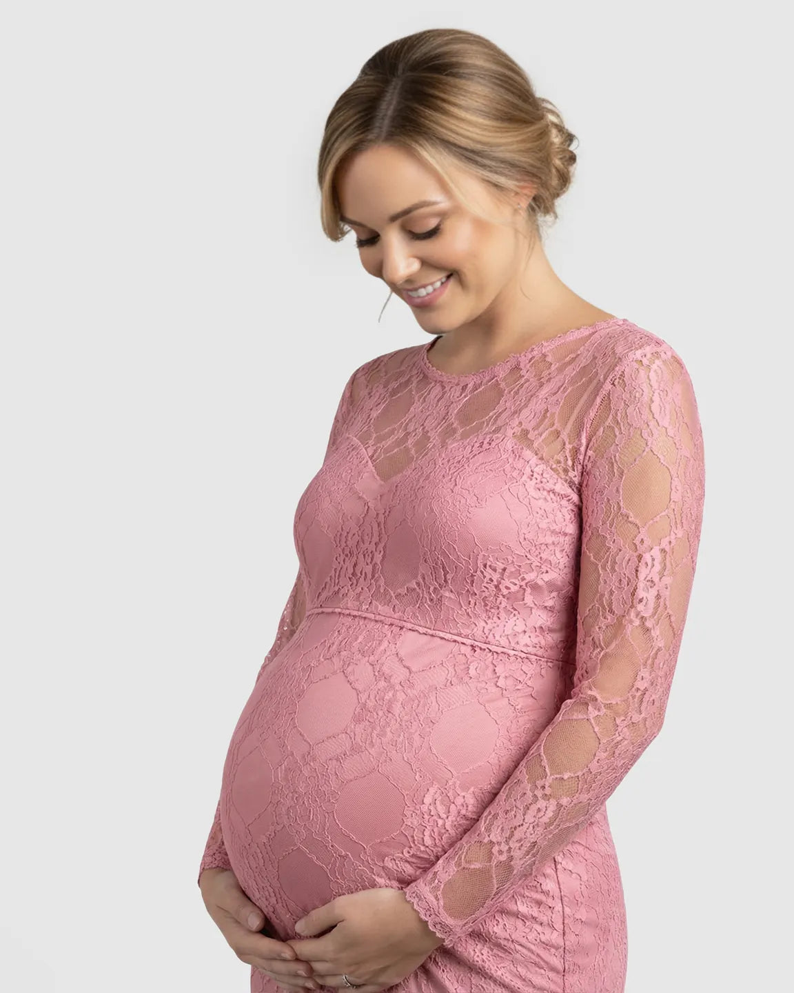 Smiling pregnant woman in a pink sheer lace long-sleeve trumpet maternity gown, cradling her bump.