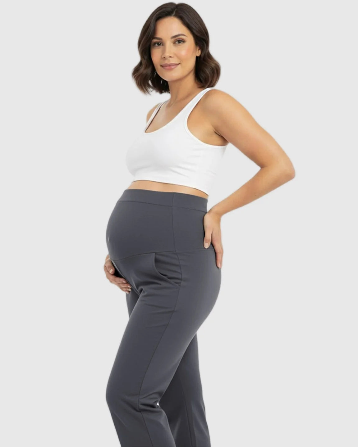 Pregnant woman in dark gray stretch flared maternity pants with pockets and a white crop top.