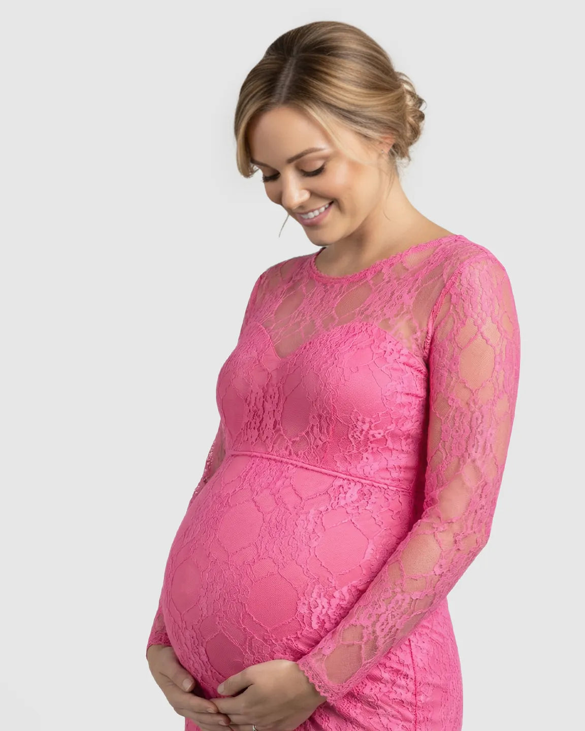 Smiling pregnant woman in a sheer pink lace long-sleeve trumpet maternity gown, cradling her bump.