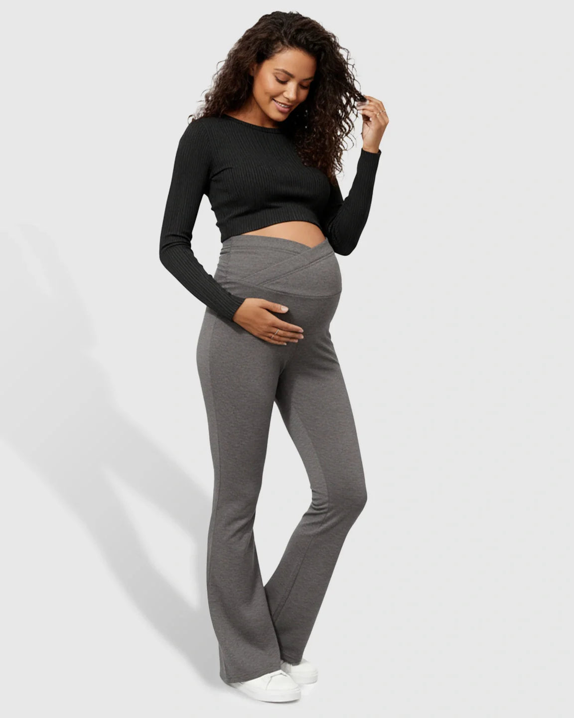 Smiling pregnant woman in charcoal gray crossed waistband flared maternity pants and a black top.