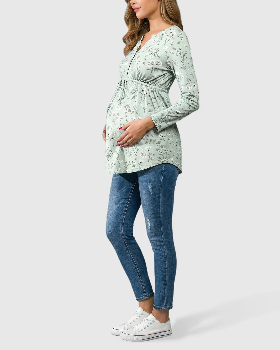 Pregnant woman in a sage green floral notch neck maternity tunic with drawstring waist, paired with jeans and white sneakers.