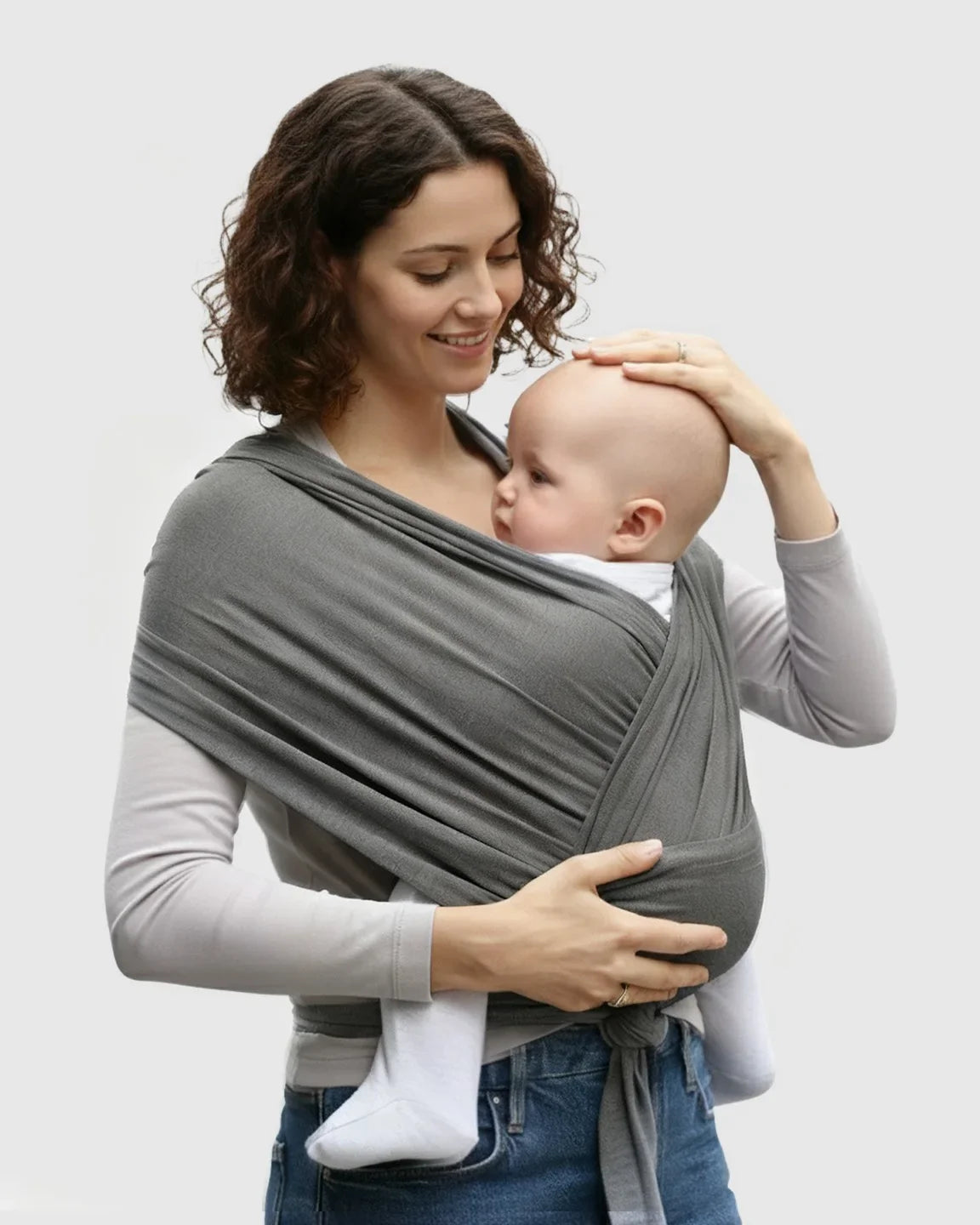 Woman smiling, wearing a dark gray adjustable wrap-around baby carrier with a baby snuggled close.
