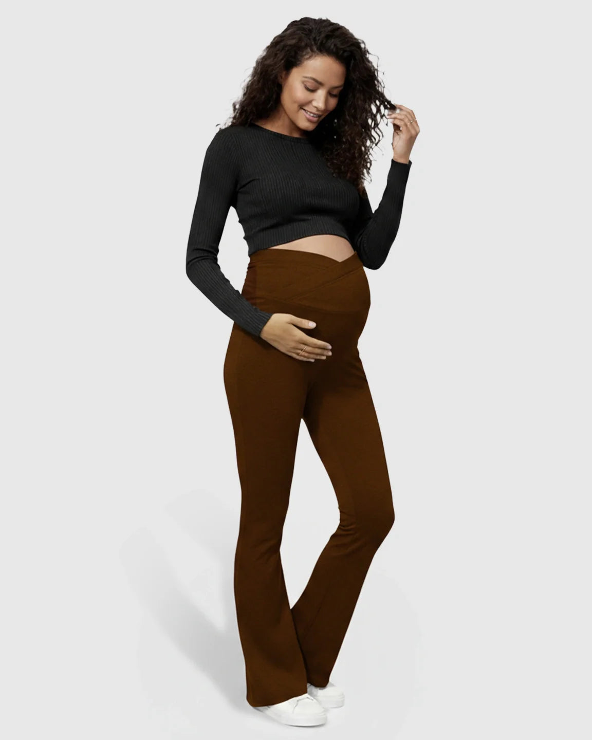 Smiling pregnant woman in brown flared maternity pants with a crossed waistband and black long-sleeve top.