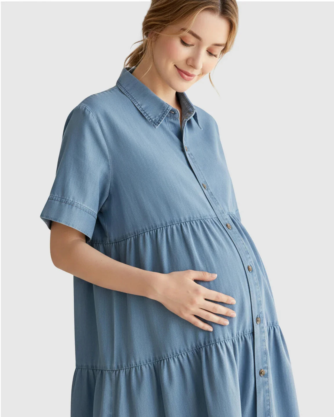 Pregnant woman in a blue tiered denim casual maternity midi dress, gently touching her belly.