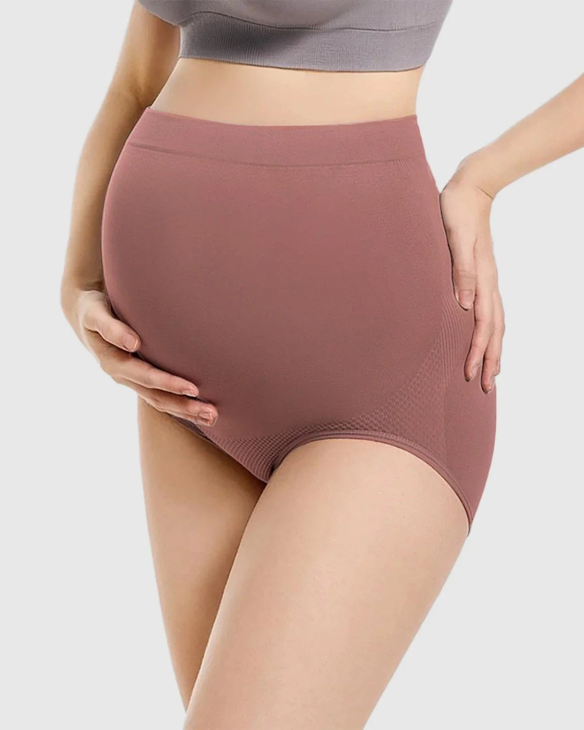 Pregnant woman wearing high-waist seamless maternity panties in a soft, dusty rose color, supporting her bump.