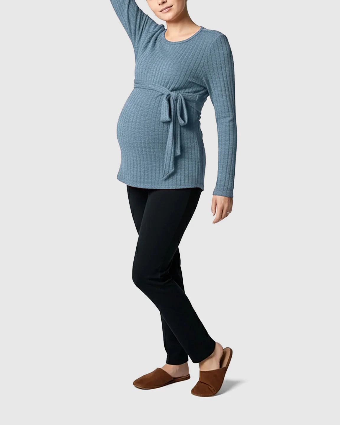 Maternity model in a blue ribbed tie-waist long sleeve sweater and black pants, with brown slippers.