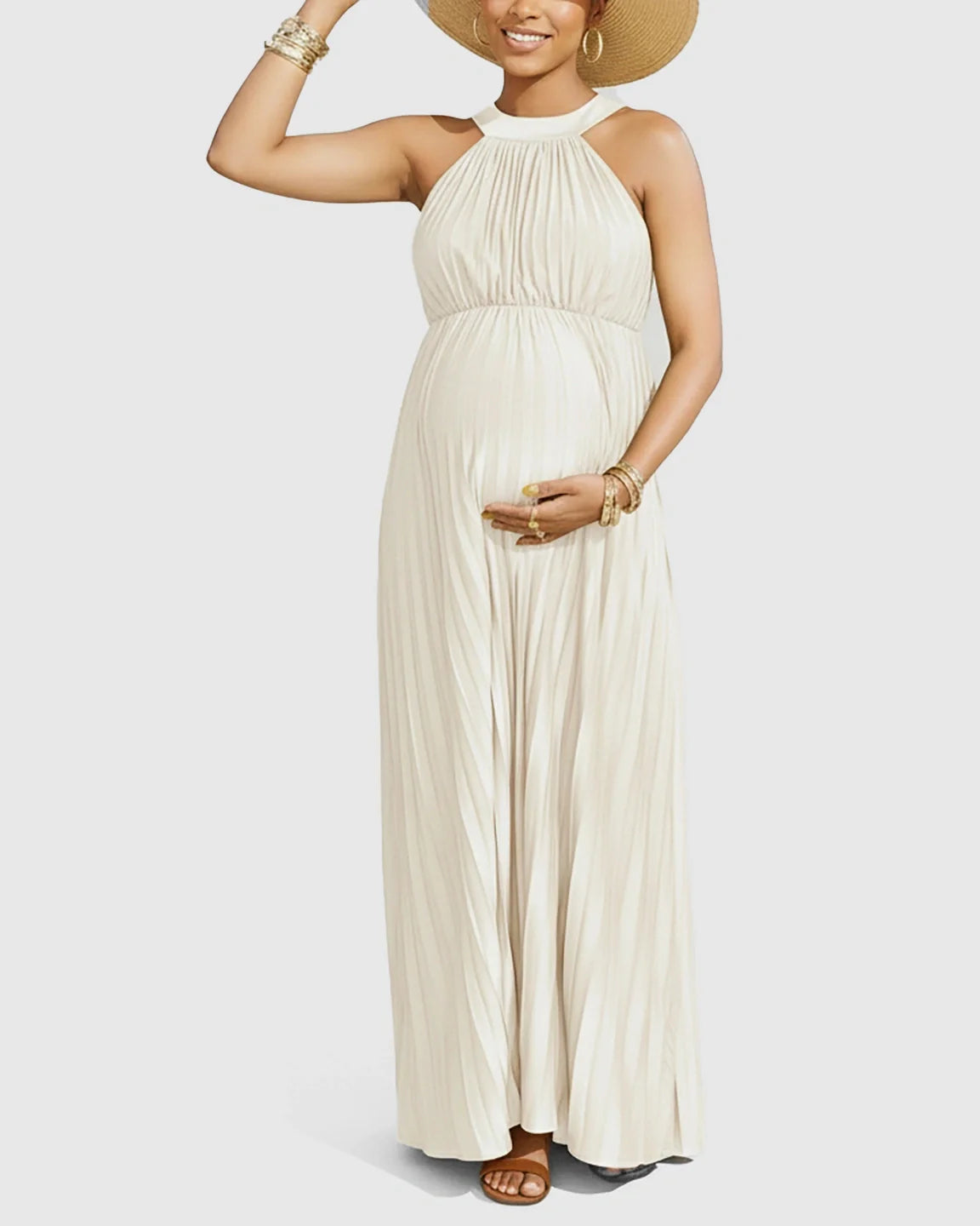 Pregnant model in a cream faux silk halter neck pleated maternity maxi dress, holding a straw hat.