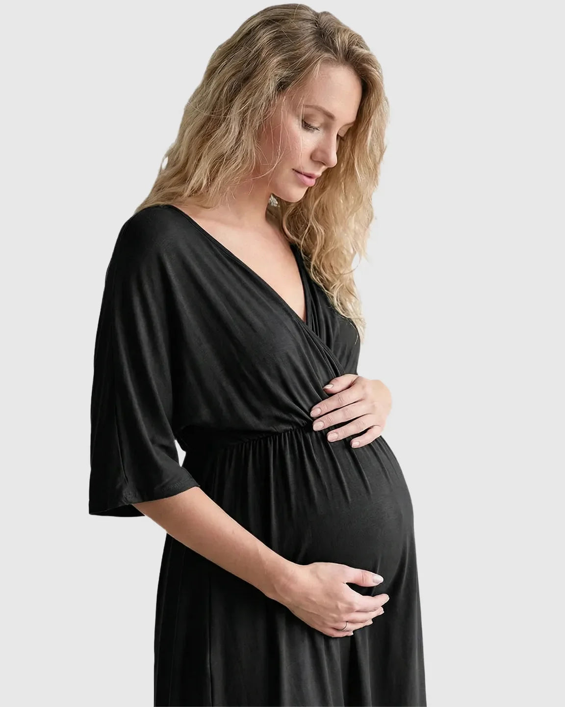Blonde pregnant woman in a black cross-front maternity midi dress with 3/4 sleeves, holding her bump.