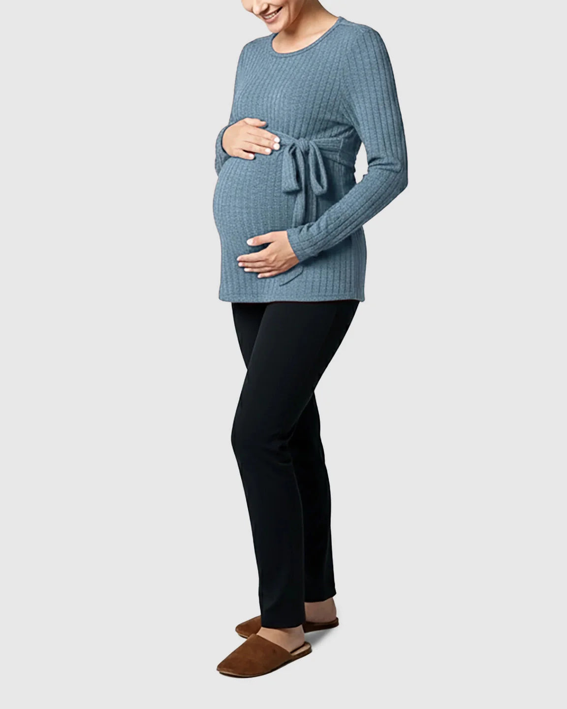 Maternity model in a blue ribbed tie-waist long sleeve sweater, black pants, and brown slippers.