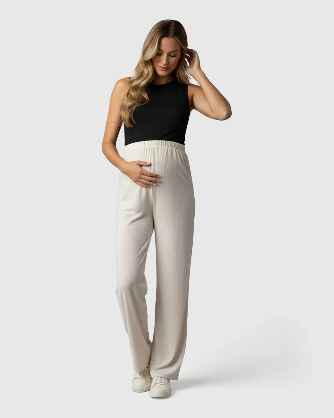 Pregnant woman in cream elastic waist wide-leg maternity pants with a black tank top, hands on belly.