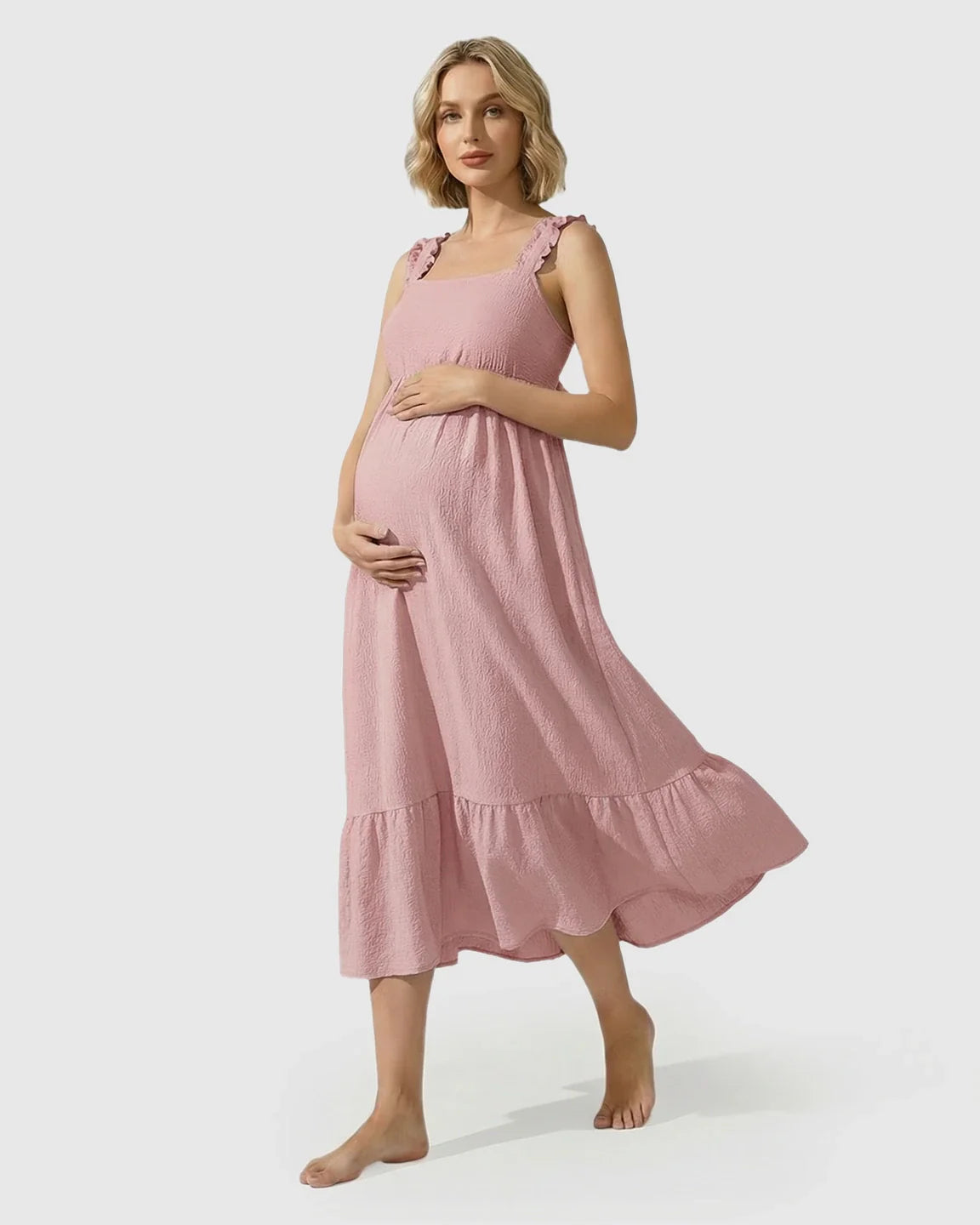 Pregnant woman in a pink ruffled strap tiered maxi dress, hands on bump.