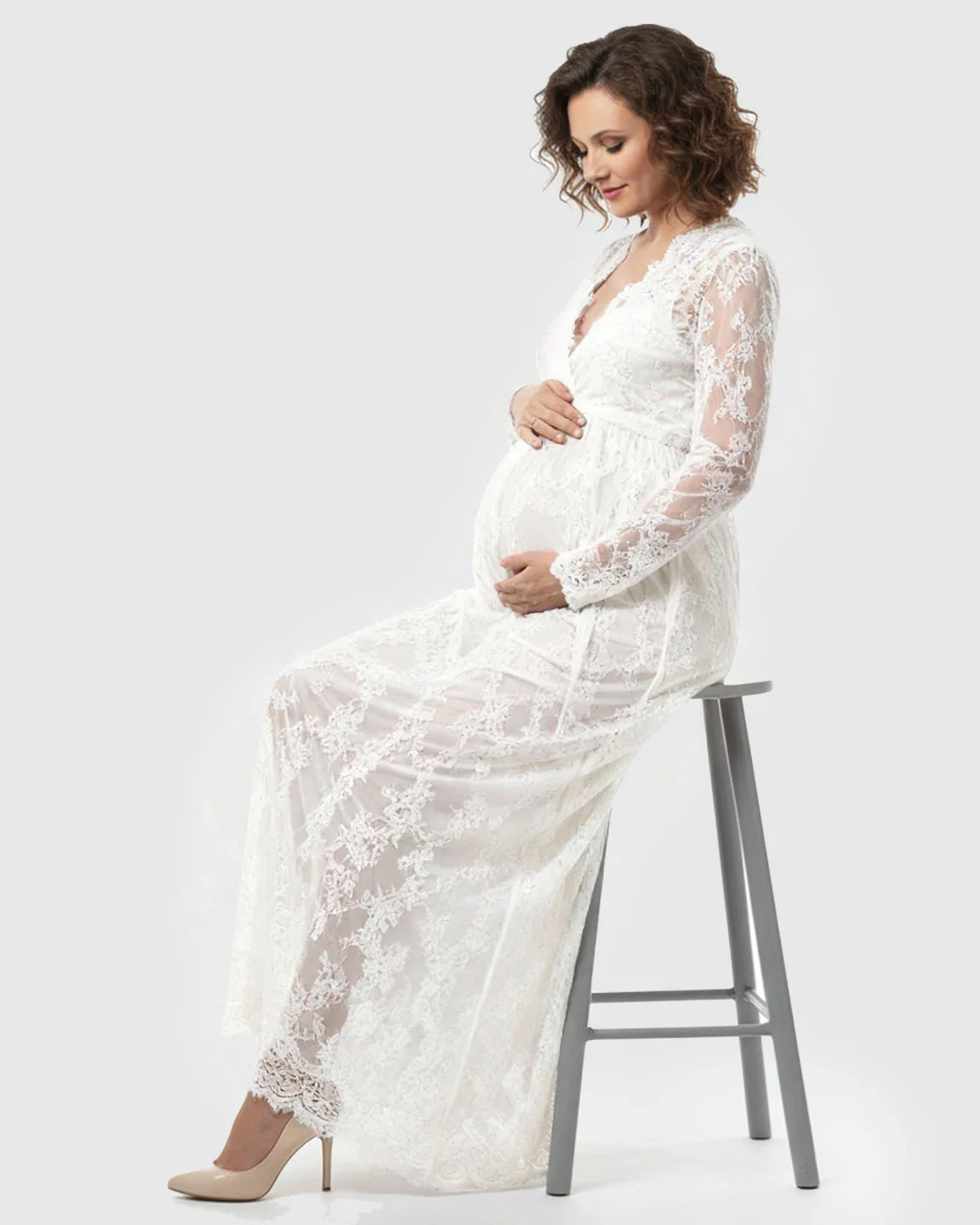 Pregnant woman in a white sheer floral lace empire waist maternity maxi dress, gently cradling her bump.