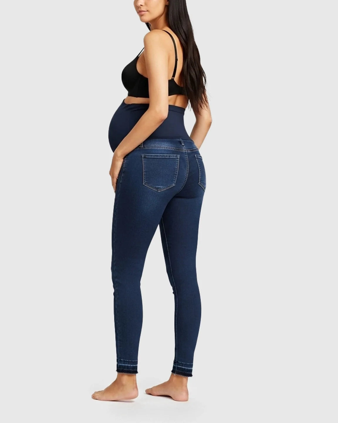 Pregnant woman from behind wearing dark wash skinny maternity jeans with a high-waist elastic panel and raw hem.