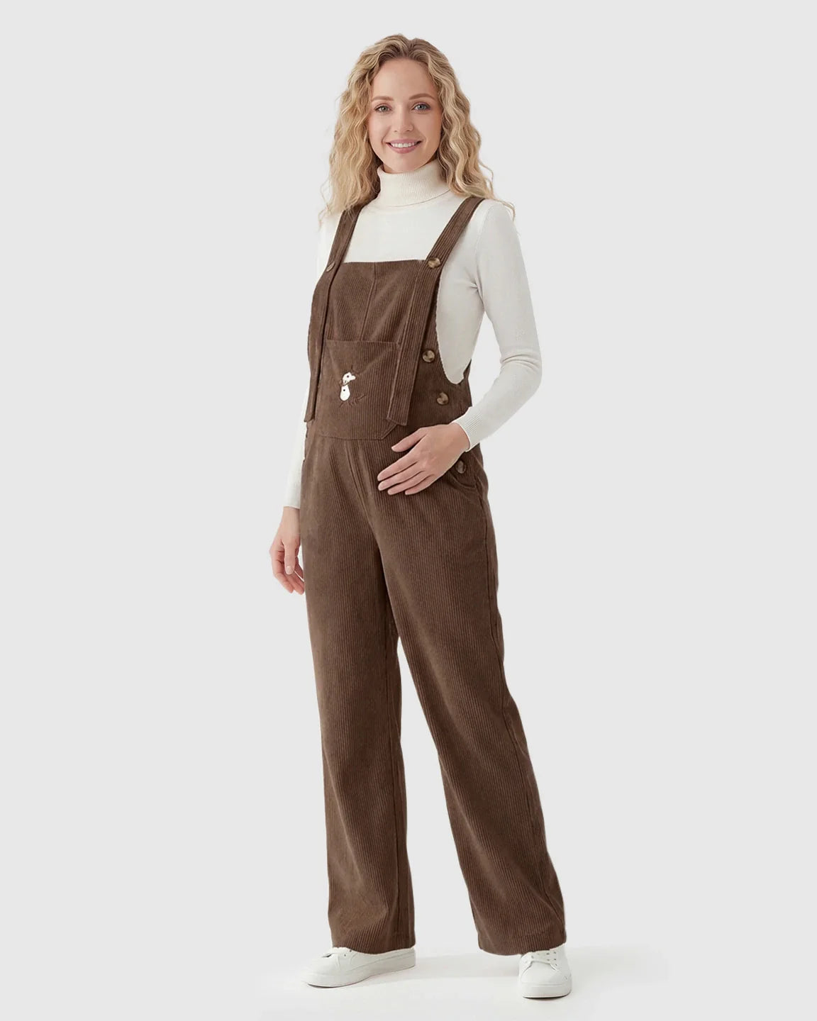 Smiling pregnant woman in brown corduroy maternity overalls with adjustable straps and a white turtleneck.