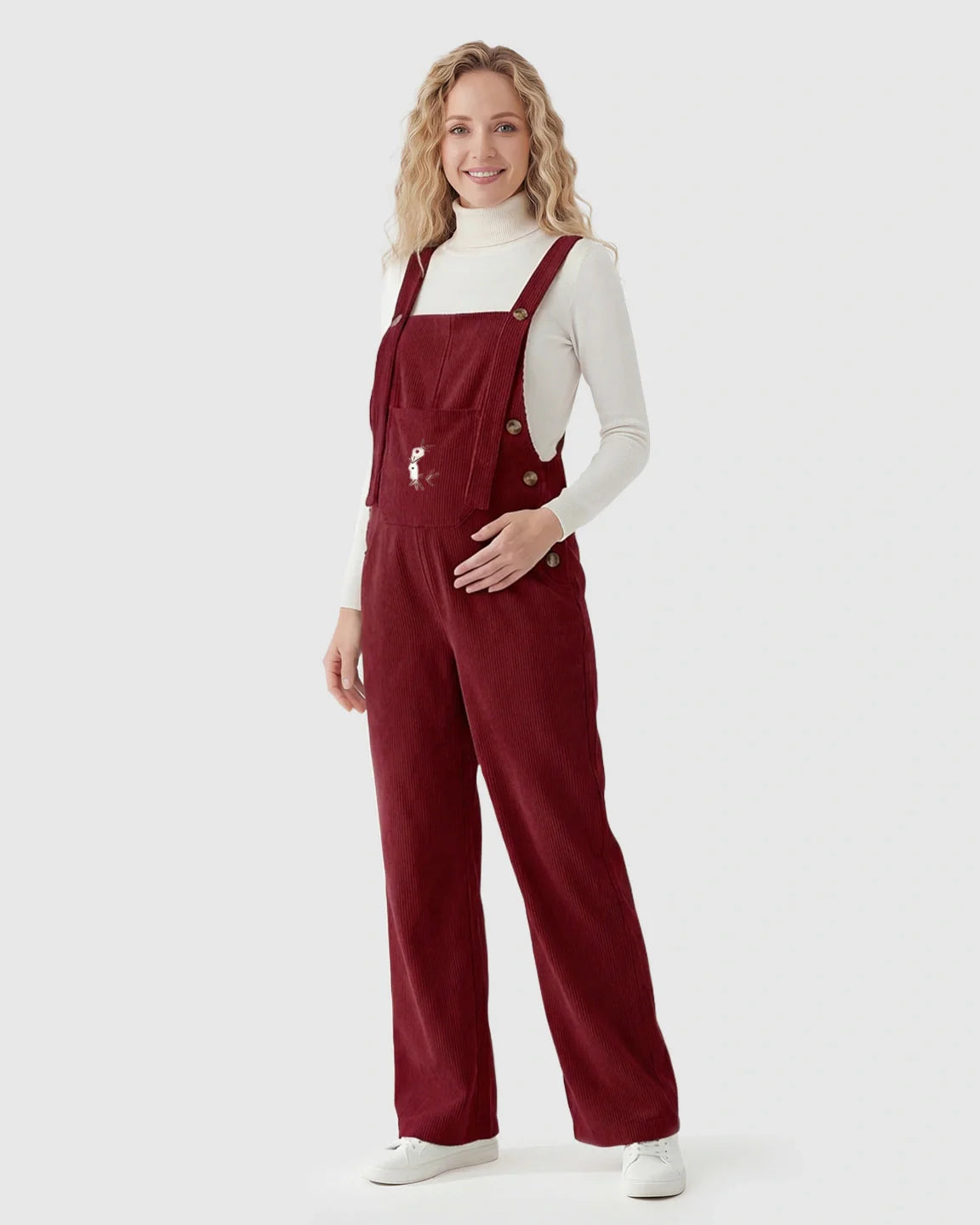 Smiling pregnant woman in red corduroy maternity overalls with adjustable straps over a white turtleneck.