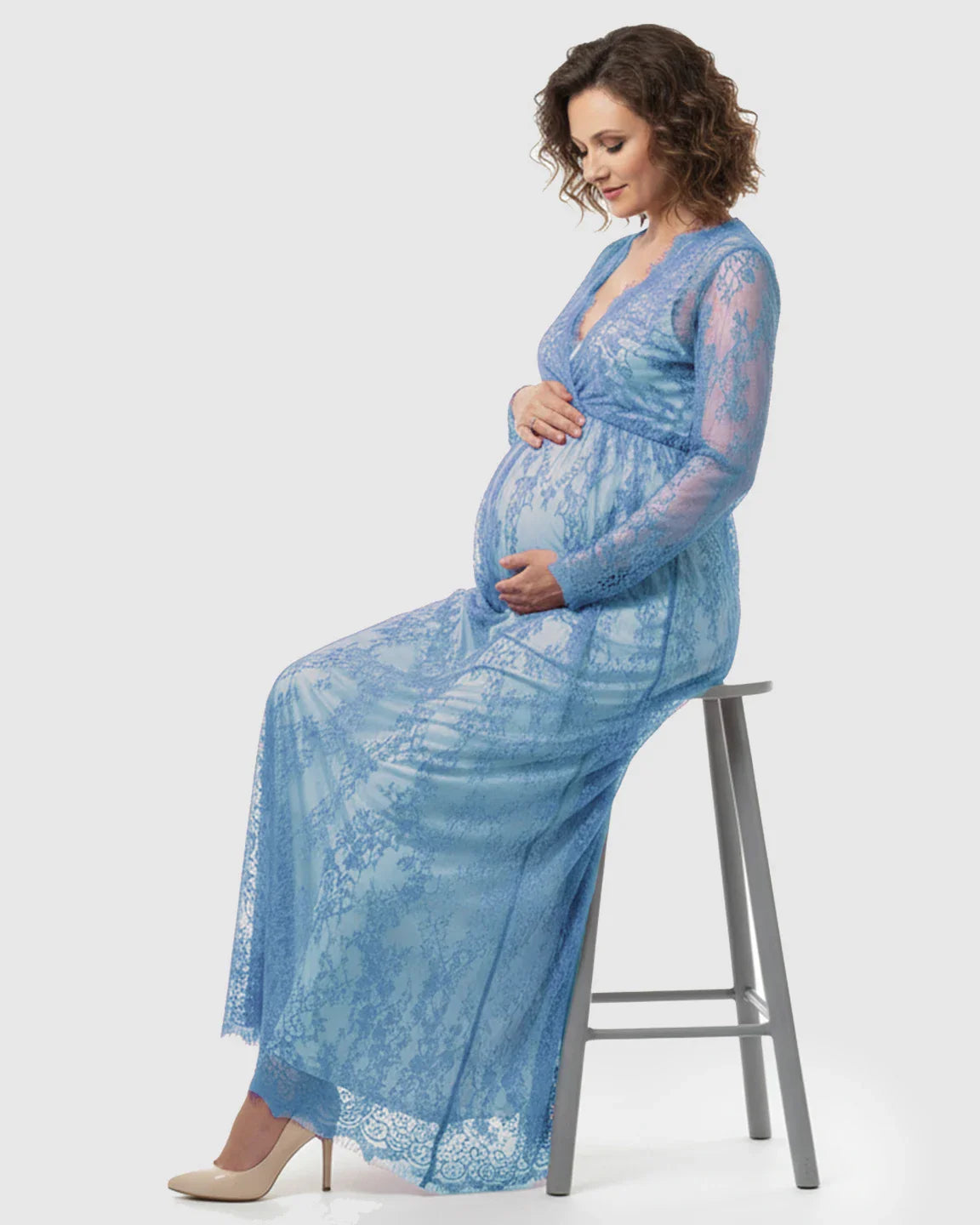Maternity model in a blue sheer floral lace empire waist maxi dress, seated and cradling her bump.