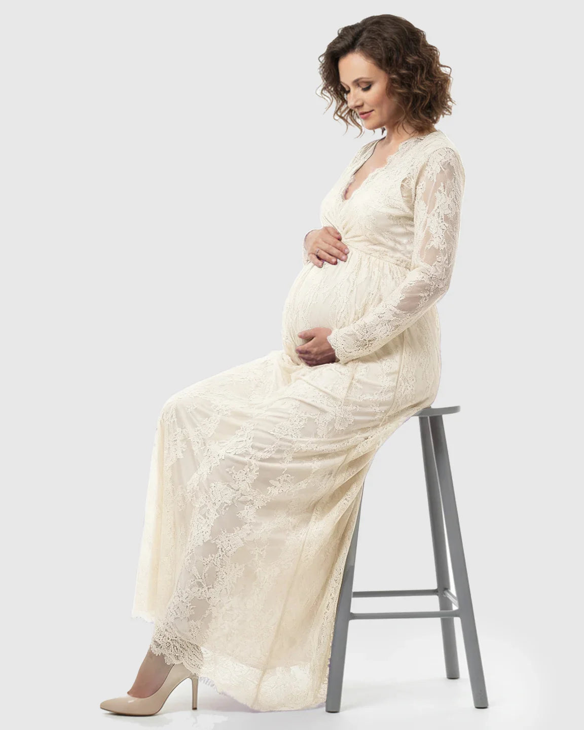 Pregnant woman in a cream sheer floral lace empire waist maternity maxi dress, gently holding her bump.