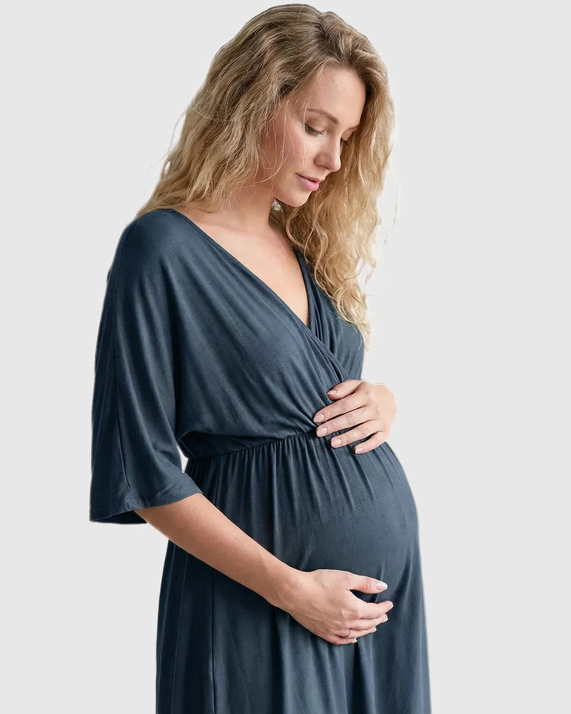 Blonde pregnant woman in a soft, dark blue cross-front maternity midi dress with 3/4 sleeves, cradling her bump.