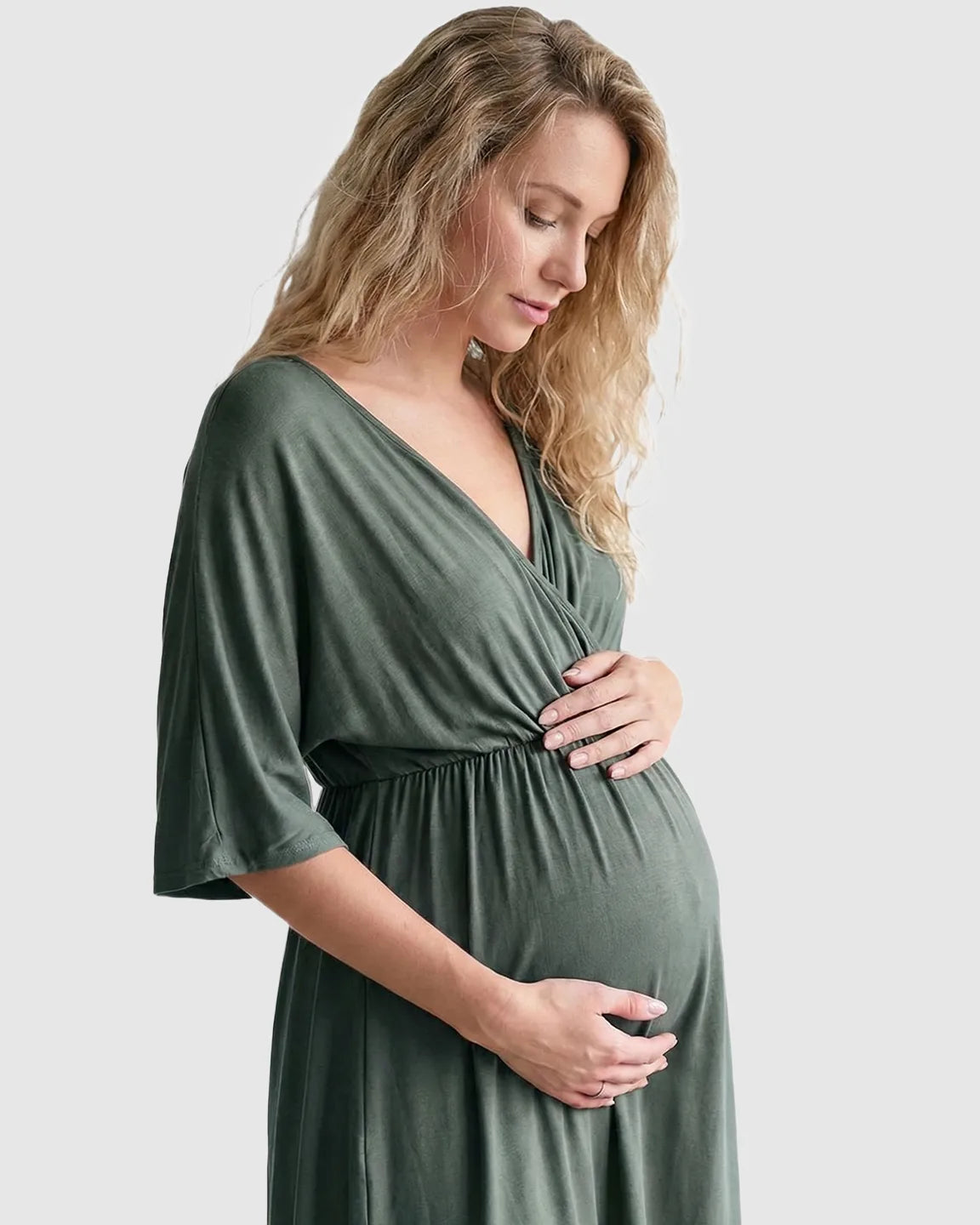Pregnant woman in a soft olive green cross-front maternity midi dress with dolman sleeves, cradling her bump.