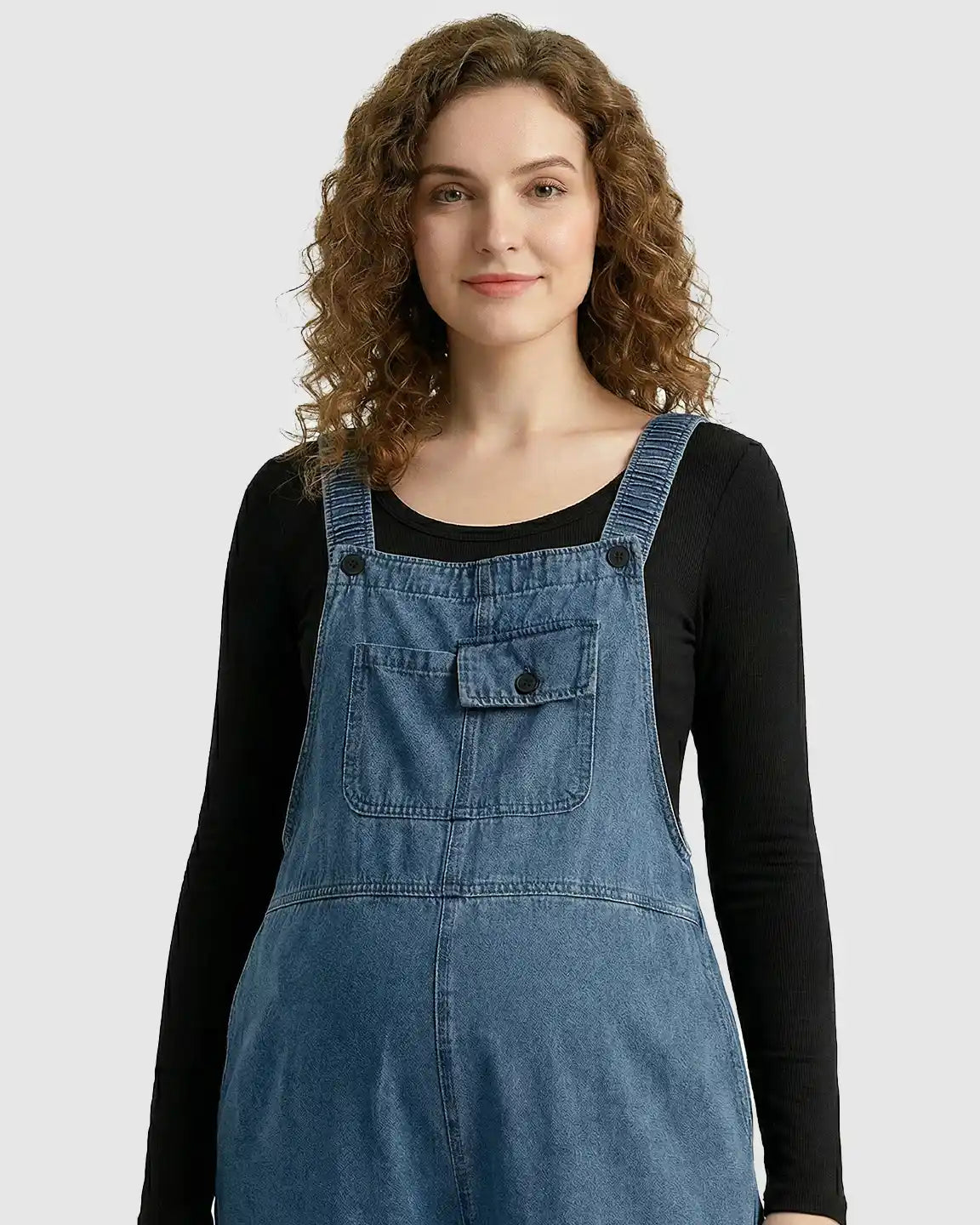 Woman in blue loose-fit denim maternity overalls with adjustable straps and front pockets over a black long-sleeve shirt.
