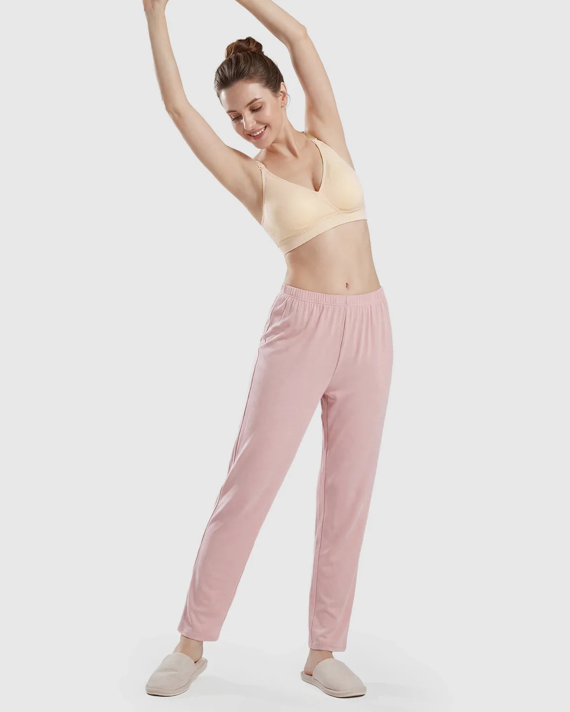 Woman in a beige seamless cross-front wireless nursing bra and pink pants, stretching comfortably.