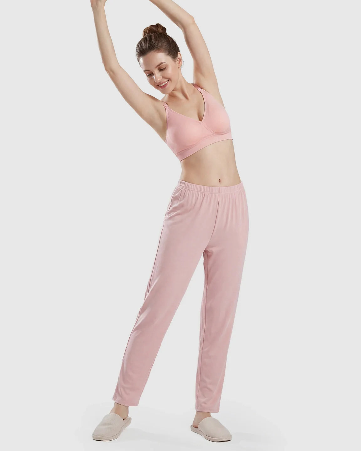 Woman in a light pink seamless cross-front wireless nursing bra and matching pants, stretching comfortably.
