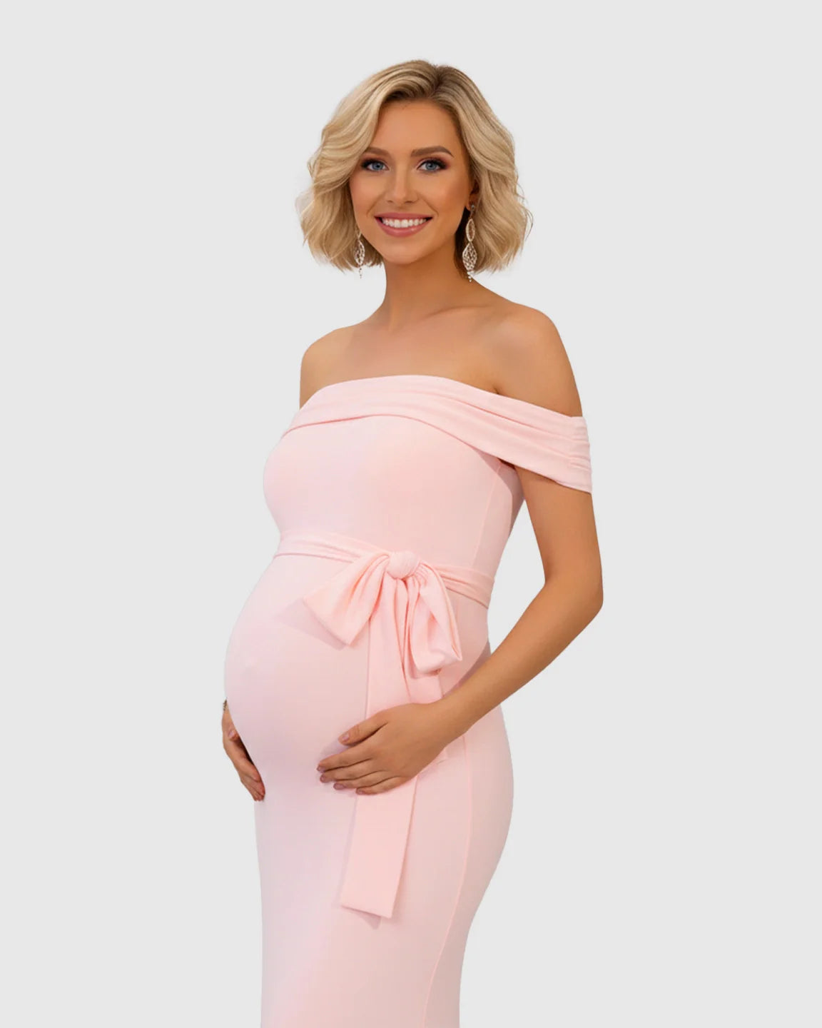 Blonde pregnant woman in a pink off-shoulder maternity gown with an adjustable tie-waist and flowing fishtail hem.