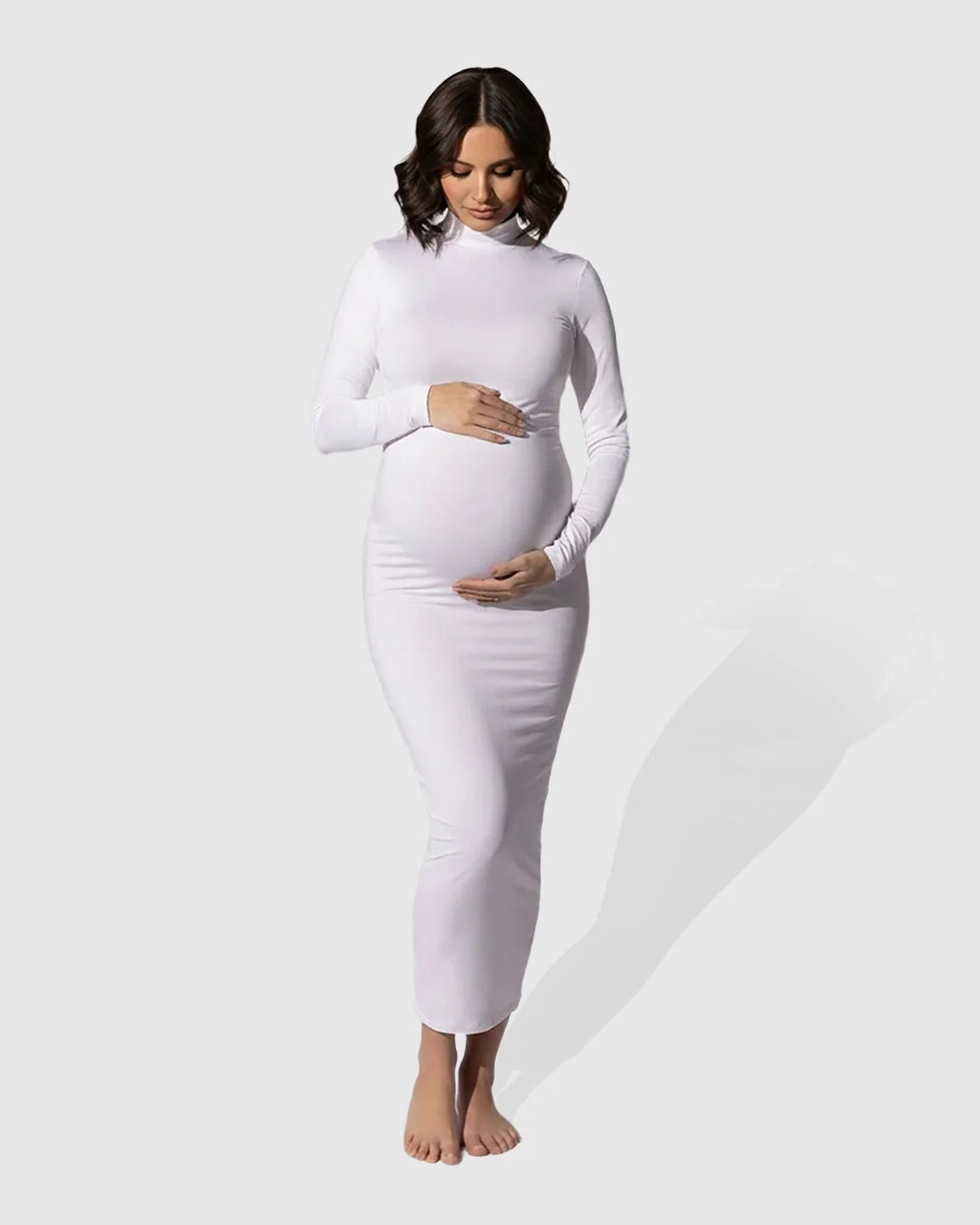 Maternity model in a white mock-neck bodycon maxi dress, long sleeves, hugging her baby bump.
