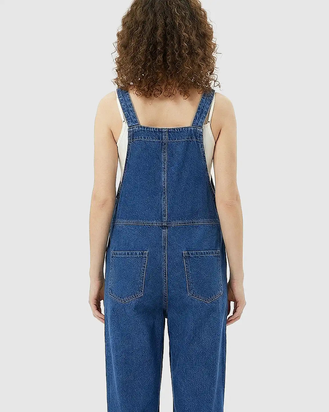 Back view of a woman wearing a loose-fit, medium-wash denim maternity jumpsuit with adjustable straps and back pockets.