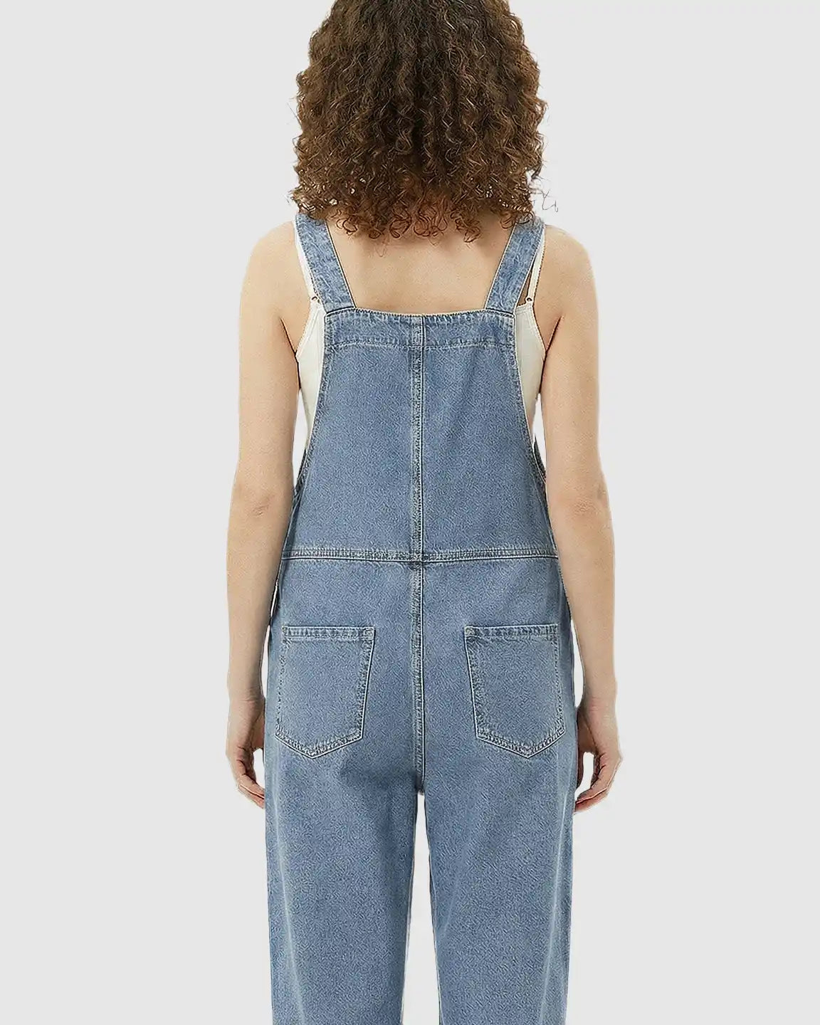 Back view of a woman wearing a loose-fit, light blue denim maternity jumpsuit with adjustable straps and back pockets.