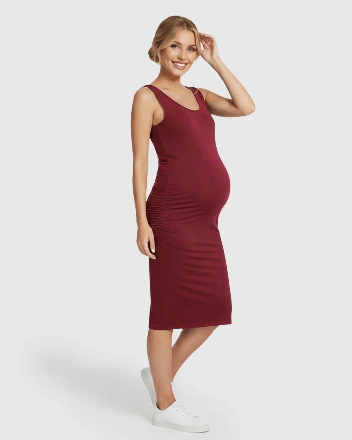 Smiling pregnant woman in a burgundy sleeveless bodycon maternity dress with side ruching and white sneakers.