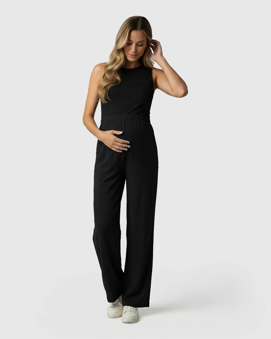 Pregnant woman in black elastic waist wide-leg maternity pants and a black sleeveless top, gently touching her belly.