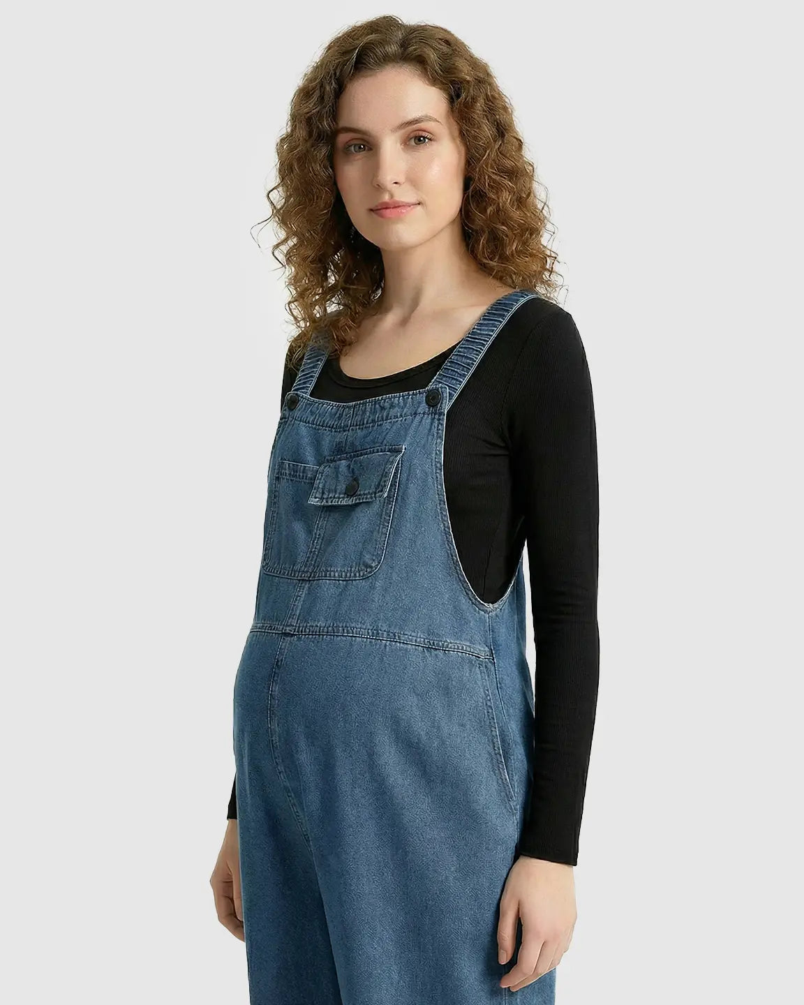 Maternity model in loose-fit blue denim overalls with adjustable straps and chest pocket, worn over a black long-sleeve top.