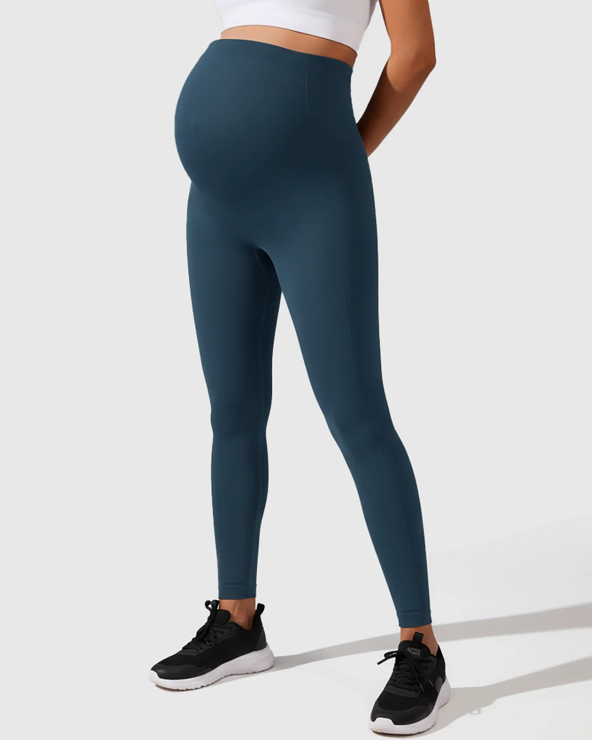 Pregnant model wearing seamless, high-waisted, dark teal maternity leggings with full-belly support.