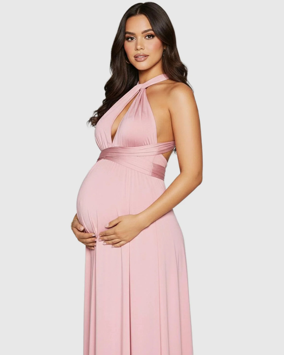 Maternity model in a pink tie-waist flowy maxi dress with a halter neckline, cradling her bump.