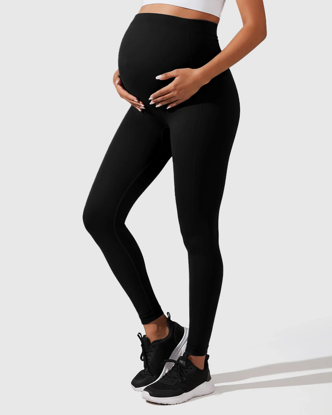 Pregnant woman in black seamless, high-waisted maternity leggings, gently supporting her bump.