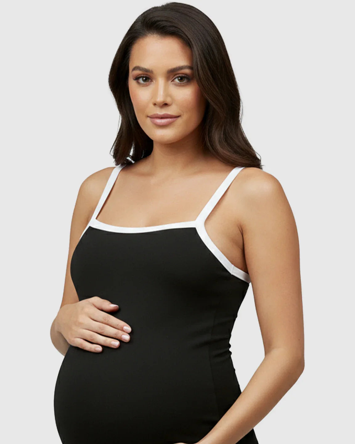 Maternity model in black sleeveless jumpsuit with white trim, holding her baby bump.