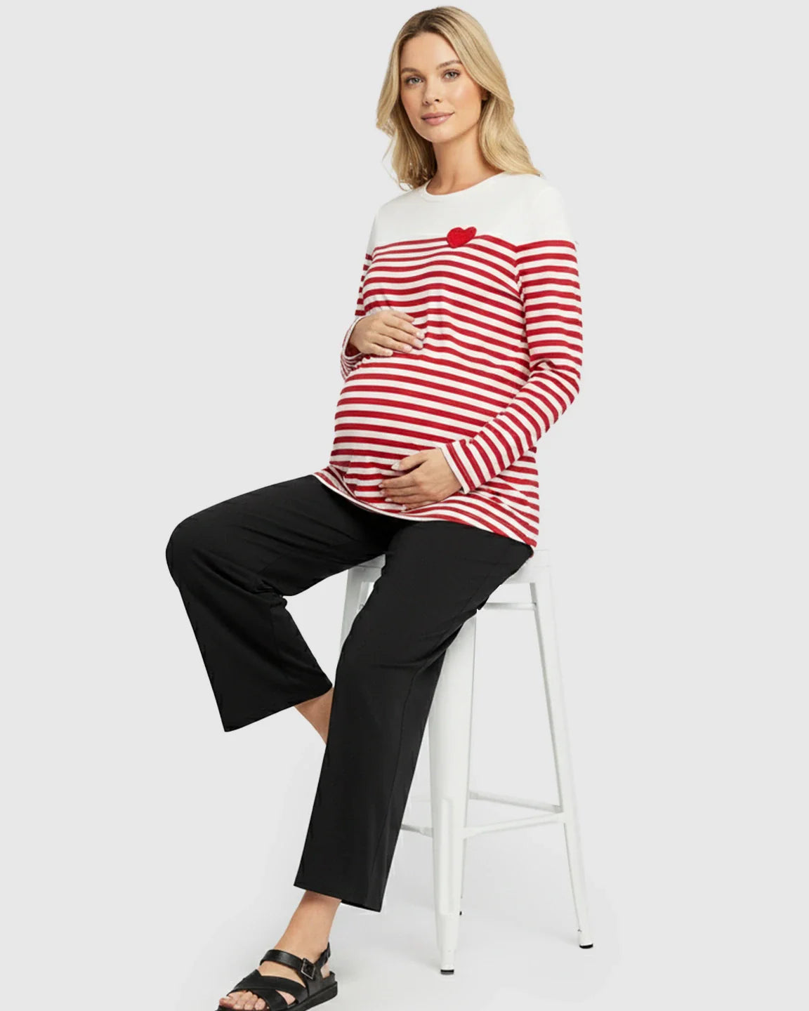 Pregnant woman in a red and white striped long-sleeve maternity top with a red heart, black pants, and sandals.