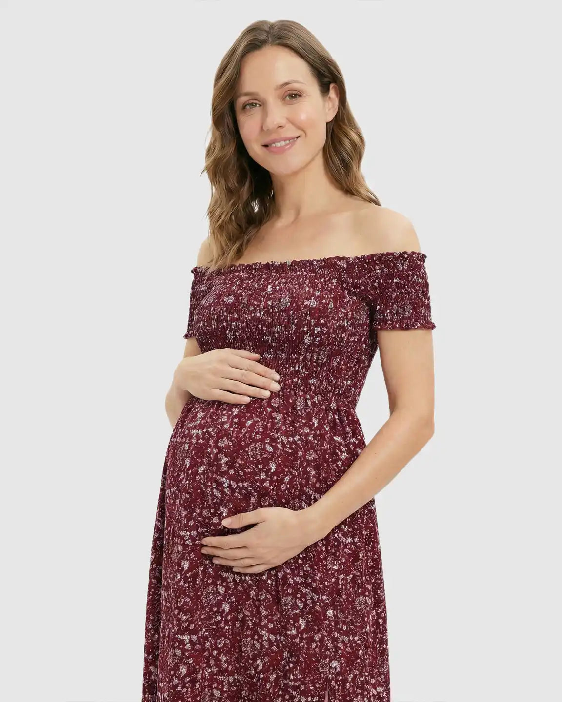 Smiling pregnant woman in a red floral off-shoulder maternity maxi dress with a side slit.