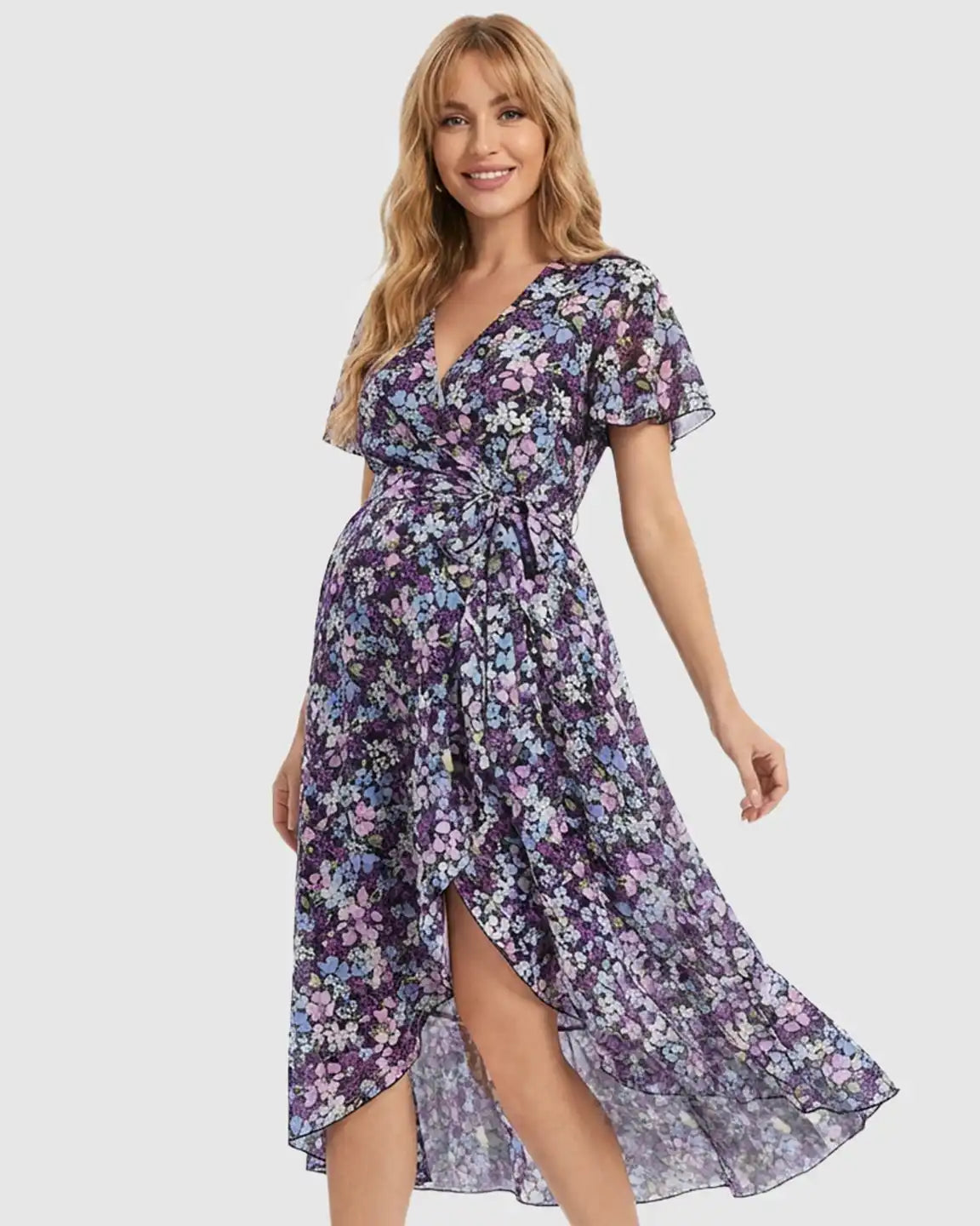 Smiling pregnant woman in a purple floral V-neck maternity midi dress with ruffled hem and tie waist.