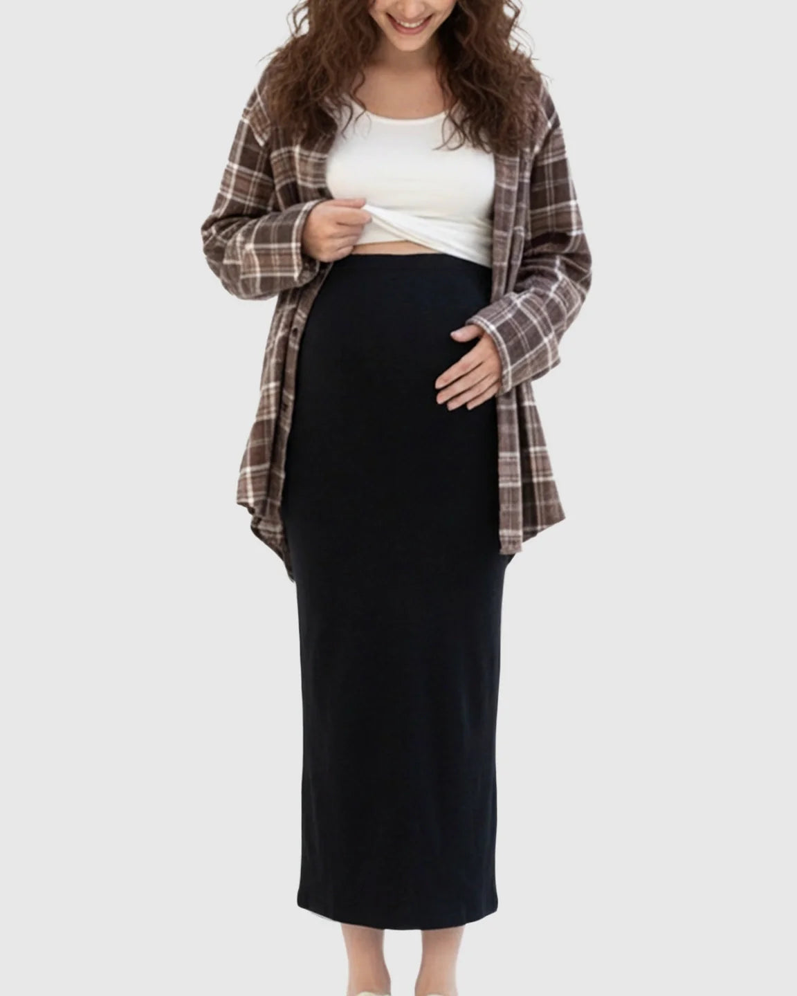 Smiling pregnant woman in a black maternity midi skirt, white top, and brown plaid shirt.