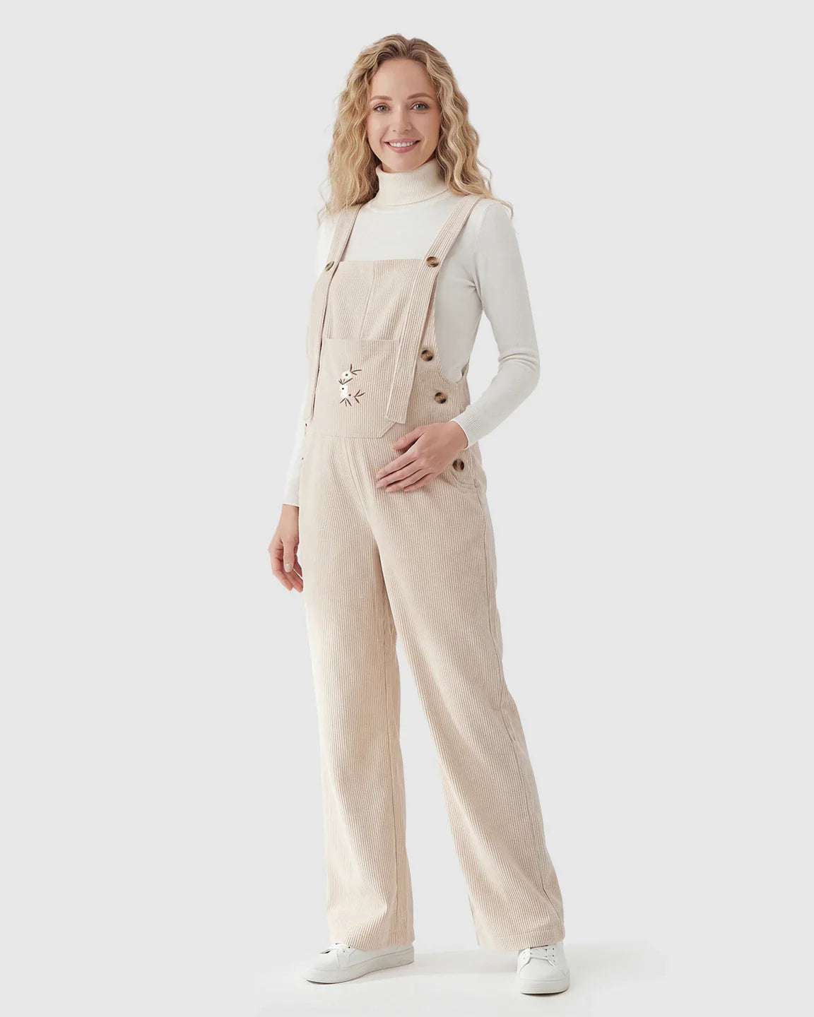 Smiling pregnant woman in beige corduroy maternity overalls with adjustable straps and wide legs over a white turtleneck.
